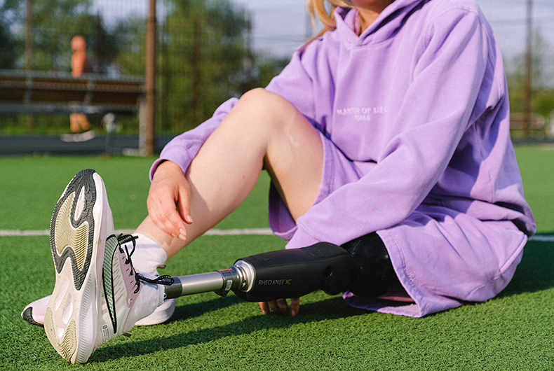 Conferência Municipal dos Direitos da Pessoa com Deficiência: garota, mostrada apenas do pescoço para baixo, sentada em um campo esportivo, utilizando um casaco roxo, com uma perna robótica estendida e a outra perna dobrada